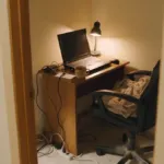 Kút spálne v noci premenený na pracovňu, starší notebook na úzkom stole pod stolovou lampou, zamotané káble a predlžovačka na koberci, pohľad od dverí
